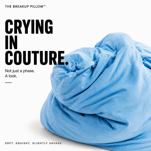 A light blue plush pillow, labeled 'THE BREAKUP PILLOW', styled as a crumpled mess on a white background with bold black text reading 'CRYING IN COUTURE.' and smaller tagline text.