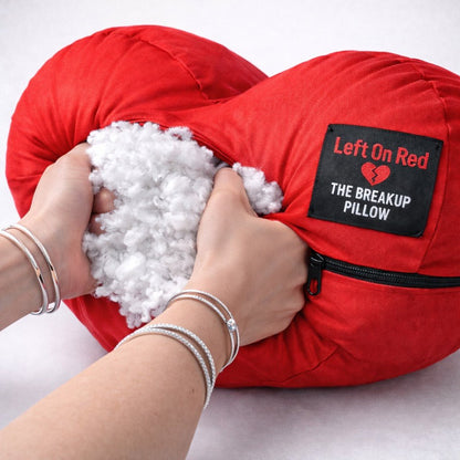 Red 'The Breakup Pillow' with a person's hands holding it open, showing white stuffing.