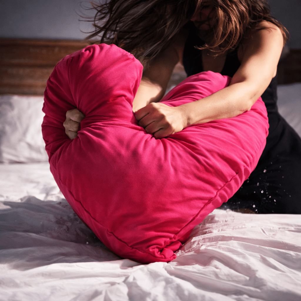 woman is throwing a large pink heart-shaped pillow on a bed.